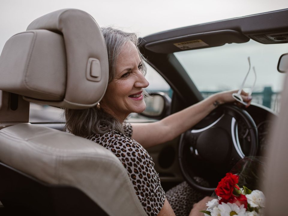 Een lachende oudere vrouw achter het stuur van haar auto.